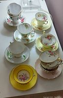 Photo showing seven footed cups and saucers with varied floral patterns, some in yellow, some in white with multiple flower colors and gold trim