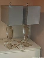 Pair of clear crystal base table lamps with silver rectangular shades placed side by side on a white surface showing overall size and style.