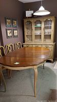 Dining table with wood top and antiqued cream base, cabriole legs, six chairs with carved details, and china cabinet in background