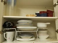 Upper cupboard shelf with stacked white plates and bowls, ceramic ramekins, small wooden bowl with spoon