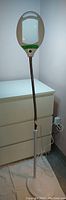 Full view of the floor lamp magnifier standing beside dresser, showing the tall white plastic body with flexible metal neck and large magnifier head with handle and button.
