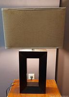 Full front view of the modern table lamp showing black rectangular hollow base and gold/taupe pattern rectangular shade in off state.
