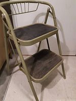 Front angle of taupe metal two-step ladder showing rubber-treaded steps and integrated handle/backrest.