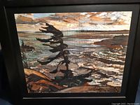Framed canvas print showing a windswept tree on a rock by water with cloudy sky, signed to represent Tom Thomson artwork