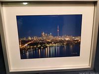 Framed night skyline print of Toronto by Norman Piluke showing city lights reflecting on water, mounted on white mat in light colored frame.