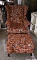 Wing chair with matching ottoman and small pillow, upholstered in French tapestry style floral fabric, orange and brown tones.