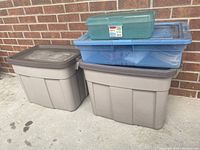 Photo showing two large gray Rubbermaid Roughneck bins stacked, with two translucent blue plastic bins and one smaller green plastic box on top, placed on concrete outside against brick wall.
