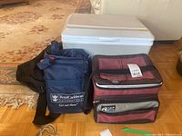 White Koolatron portable cooler with lid closed, placed behind three soft travel and cooler bags on carpet