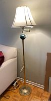 Full view of floor lamp showing the brass-colored base, green floral accent on the pole below the fabric shade, standing next to couch and wall.