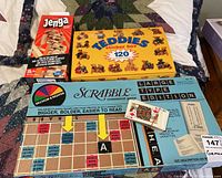 Photo showing Scrabble Large Type Edition board game box with cards, Classic Jenga game box, Teddies Sticker Box with 120 stickers and a deck of playing cards on a bed.