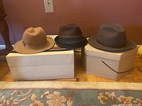 Three hats displayed on top of two cardboard hat boxes on a wooden floor with a wall background.