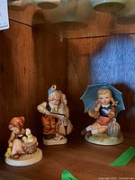 Photo of three ceramic figurines on wooden shelf including girl with umbrella, boy with cello, and girl with basket and animals