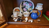 Wide shot showing entire lot including large floral plate, Nippon covered dish, blue jug, salt and pepper shakers, small vase, and other small porcelain pieces on wooden surface