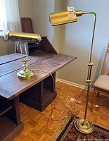 Shows both lamps together on a wooden desk and floor, exhibiting brass-colored metal finish with flexible arms and lamp heads, cords visible.