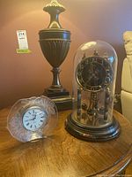 Two mantle clocks on a wooden table: smaller frosted glass clock with etched rose design and larger anniversary clock under glass dome