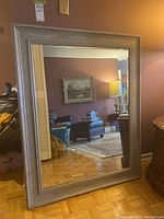 Front view of large framed mirror, showing clear reflective glass and a wide, light brown/gray frame with subtle texture. Reflections include a living room area with furniture and lamps.