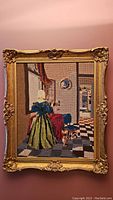 Full view of the framed needlepoint artwork showing the woman in green and blue period dress in a domestic scene, hung on a wall.