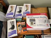 Photo showing four boxed Sigvaris compression wraps, Omron Premium Blood Pressure Monitor box, and Cura-Heat patch box on wood surface.