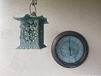 Photo showing the hanging metal candle holder with intricate floral design and pagoda-style curved roof next to the round thermometer.