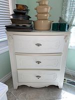 Front view of white dresser with three drawers and metal pulls, decorative carvings on drawer fronts. Various kitchen items on top not included.