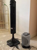 Photo of the Inertek black floor fan and the Black & Decker gray space heater side by side on a beige carpeted floor next to white wall and metal shelving.