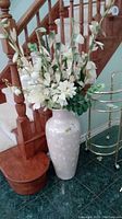 Full arrangement of faux white flowers in ceramic vase placed on green tiled floor beside wooden staircase and metal cart.