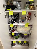 Open white closet showing five shelves with various labeled household items including a cordless phone, tapes, sealant, light bulbs, batteries, and small tools