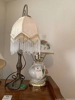 Two brass-colored lamps side by side: one with white fabric drop shade and beaded fringe, and one with white floral glass body and floral glass shade.