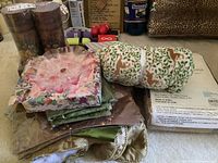 Display of folded assorted linens including tablecloths, towels, napkins in various floral and patterned fabrics along with a rose-colored decorative plate in packaging, and decorative storage boxes with animal print.
