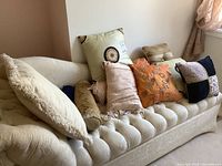 View of assortment of decorative pillows resting on a cream tufted couch including large beige, gold damask, orange with butterfly print, animal print, and black pillows.
