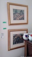 Two framed prints on a wall, one above the other, showing landscape scenes in gold frames.