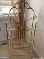 Front view of the gold-colored metal shelving unit with three folding wire shelves showing arching frame and grid shelf surfaces.