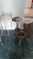 Multiple tables and plant stands including metal patio table, wooden plant stands, and metal and glass plant stand in corner of room