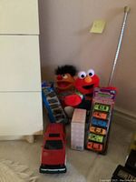 Overview photo showing Elmo and Ernie plush dolls, two toy car packages, a red remote control car, and photo album boxes stacked near a dresser.
