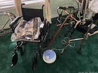 Wide view of Drive wheelchair with blankets on seat, Aingo walker, 2 canes, and Life humidifier on green carpet near window with white curtains