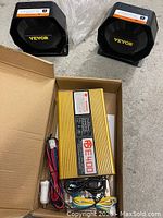 Two black horn speakers branded Vevor positioned next to an open box containing the gold-colored amplifier unit and wiring cables on a concrete floor.