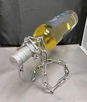 Front view of the metallic chain holder balancing a wine bottle by the neck, placed on a fabric surface against a dark backdrop.