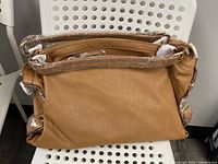 Brown classic satchel shown unwrapped on a chair, highlighting the double handles and smooth vegan PU material texture.