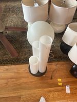 Photo shows two lamps on wooden floor. One lamp with three white tubular tops and black circular base, the tallest tube about 29 inches high. Other lamp with large white spherical top and reflective chrome base.