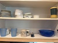Shelves with white ceramic bowls, ramekins, blue glass tumblers, mugs, and blue glass serving bowl.