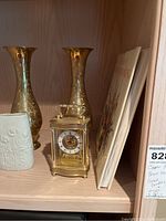 Tall copper vases, white ceramic vase, Bulova carriage clock, and two books on a wooden shelf