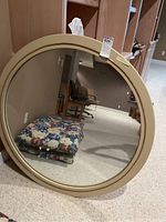 Full view of large round mirror with beige frame placed on carpeted floor showing its size and reflection.