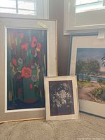 View of three framed artworks, including one large colorful floral painting, one smaller floral painting, and one landscape painting in frames.