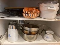 Shelf with metal pots stacked, two white CorningWare carafes with blue floral design, three copper molds, and two white plastic ice cream maker containers