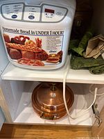 White Oster bread maker with front control panel and pictures of bread types on top shelf. Next to green kitchen cloths.