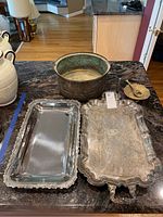 Six metal items including two silverplate trays, copper pot, brass bowl, and brass bottle opener displayed on a dark countertop.