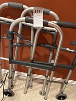 Two similar aluminum adult walkers with padded gray hand grips and front wheels seen leaning against a reddish wall.