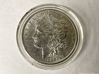 Obverse side of the 1889 Morgan Silver Dollar showing Lady Liberty in profile with date and inscriptions. Coin is encased in clear plastic holder.