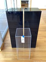 Photo shows two large black acrylic rectangular display pillars side by side with a smaller clear acrylic pillar in front of them on wood floor by glass window, highlighting size difference and glossy surfaces.