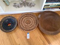 Full view of all three items placed on floor against shelf: black and beige ceramic bowl with colorful center, woven tray, and wooden bowl.